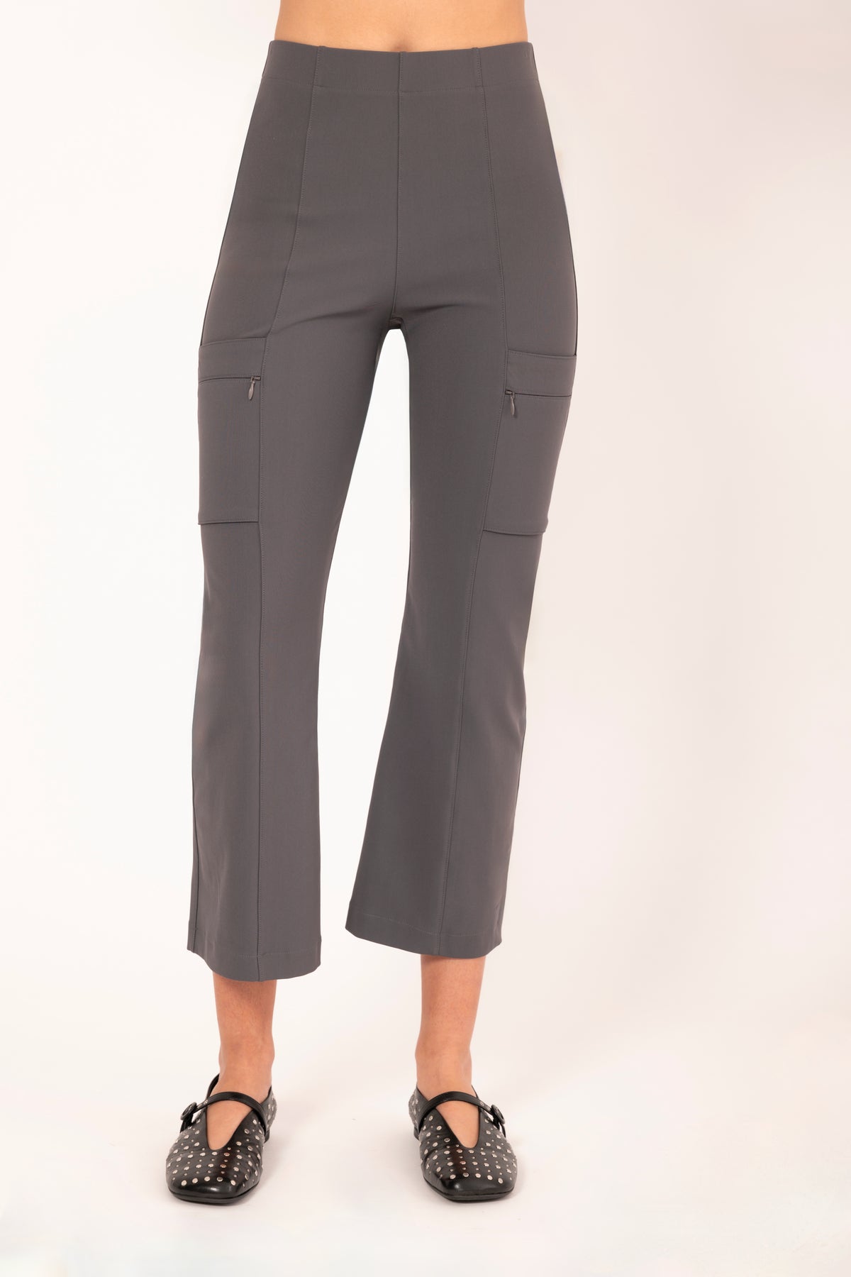 A model is shown from the waist down wearing high-waisted, cropped gray pants with side cargo pockets and black studded Mary Jane shoes, set against a plain off-white studio background.