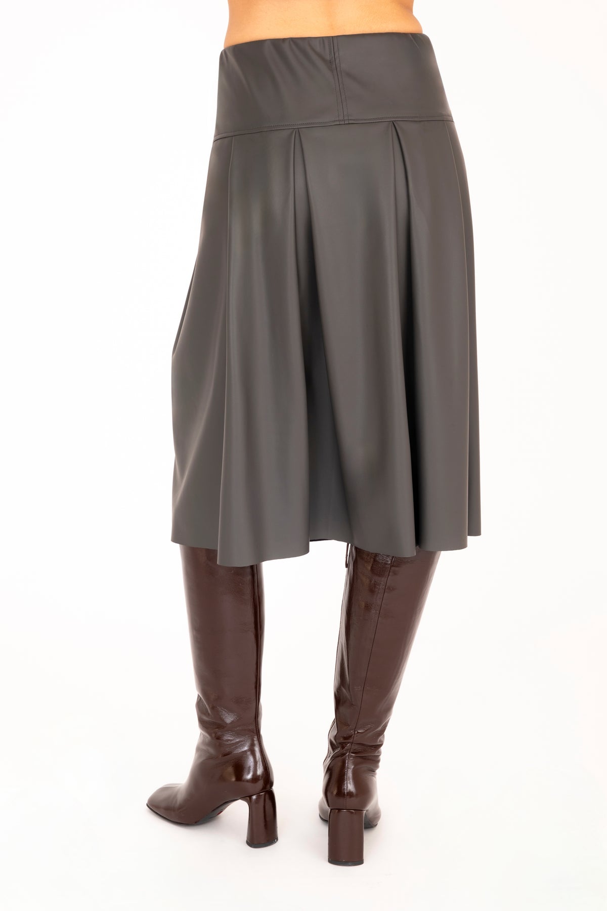 Back view of a gray vegan leather pleated A-line skirt shown on a model from the waist down, paired with dark brown knee-high leather boots against a plain white background.