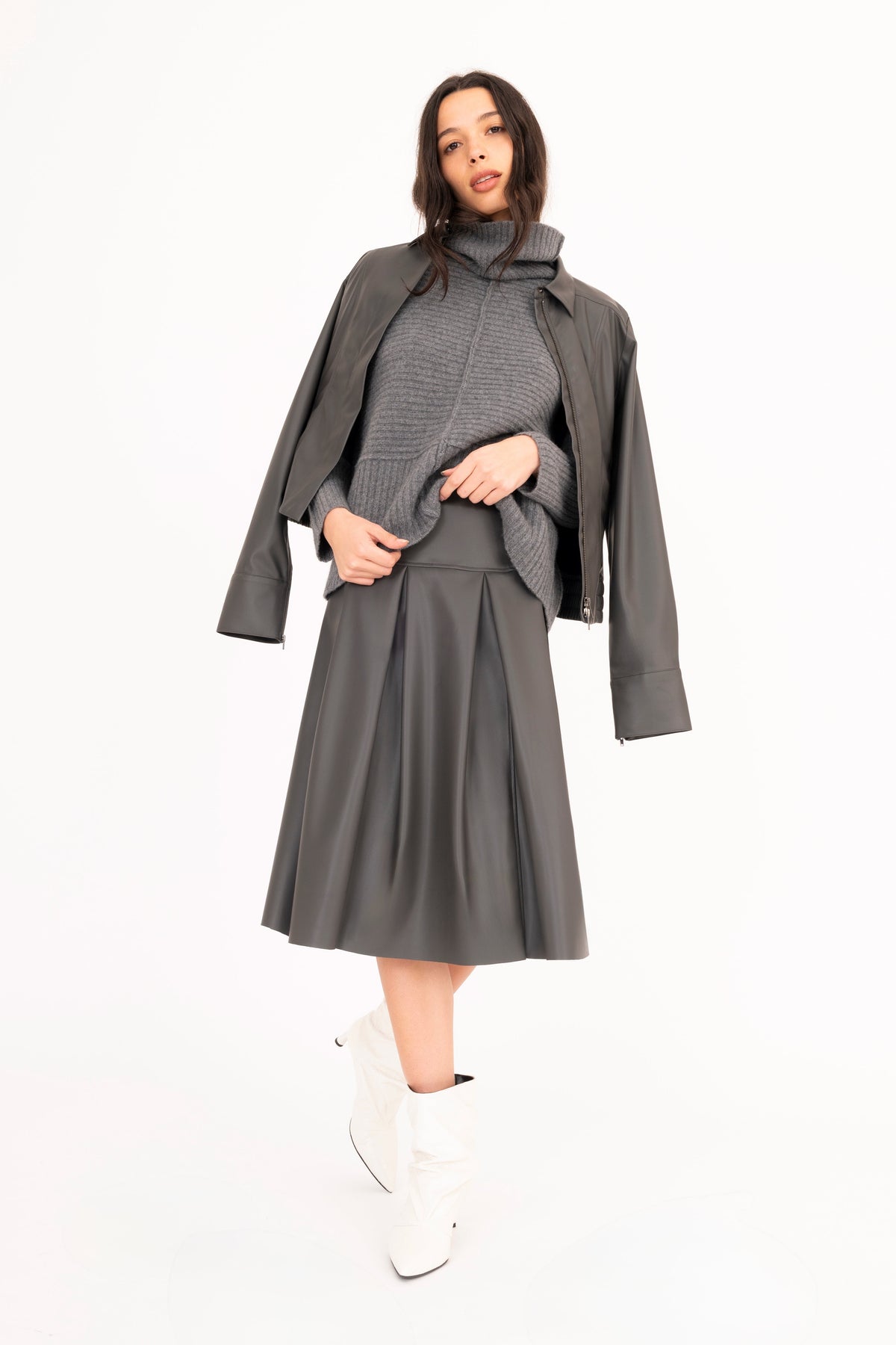 A woman wearing a gray vegan leather pleated skirt with a matching vegan leather jacket and cashmere funnel-neck sweater on a white background