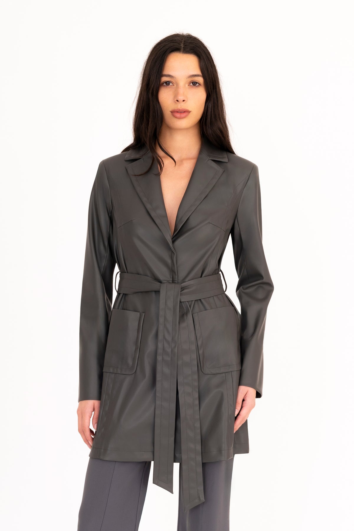 A woman modeling a dark gray vegan leather trench coat with a collar, waist tie, and front pockets, paired with matching dark gray pants, against a plain white studio background.