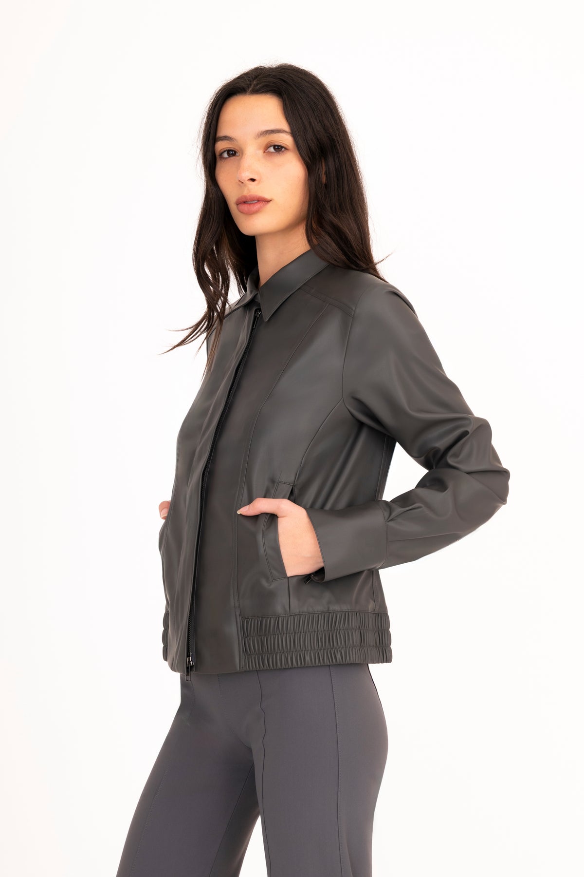 A woman modeling a dark gray vegan leather jacket with a collar, front zipper, and side pockets, paired with matching gray pants, against a plain white studio background.