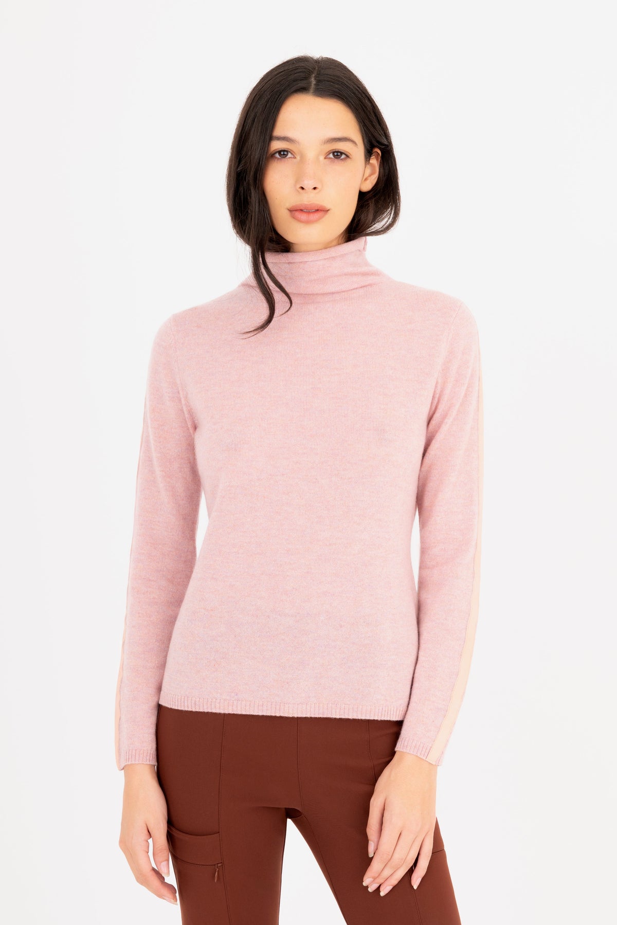 A woman modeling a light pink turtleneck sweater, paired with brown pants against a plain white studio background.