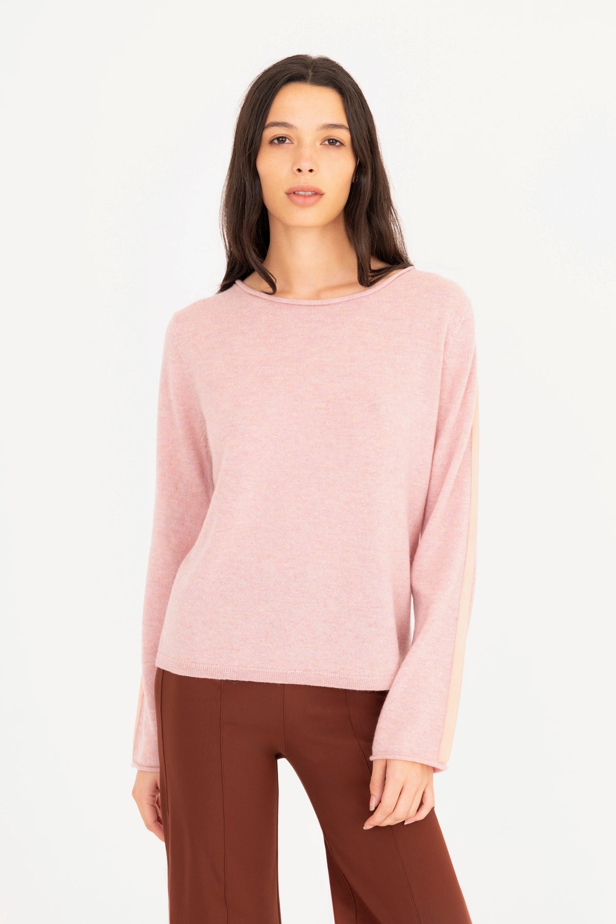 A woman modeling a pink long-sleeve knit sweater, paired with brown pants, against a plain white studio background.