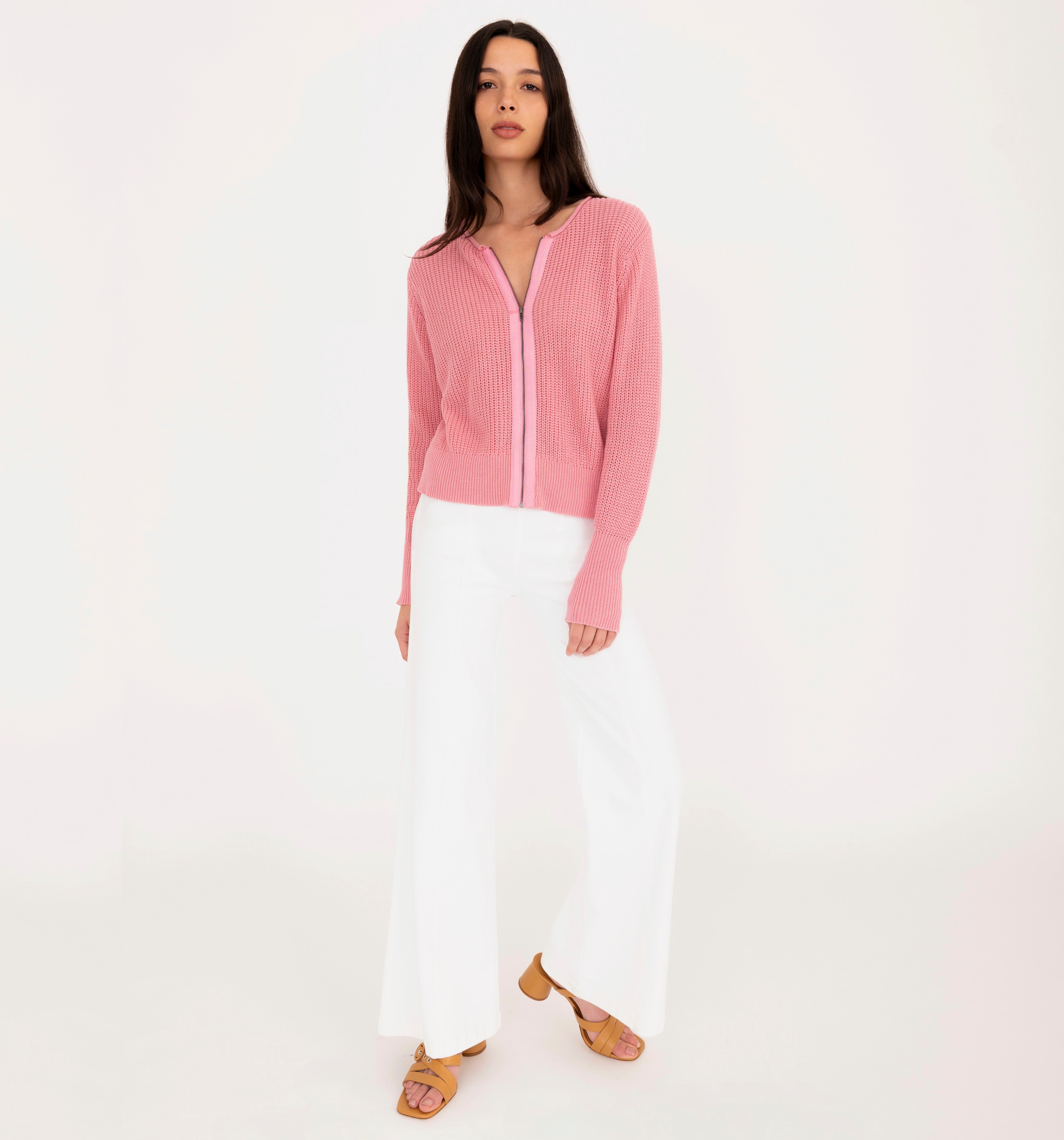 Woman wearing a pink zip-up sweater and white pants on a white background