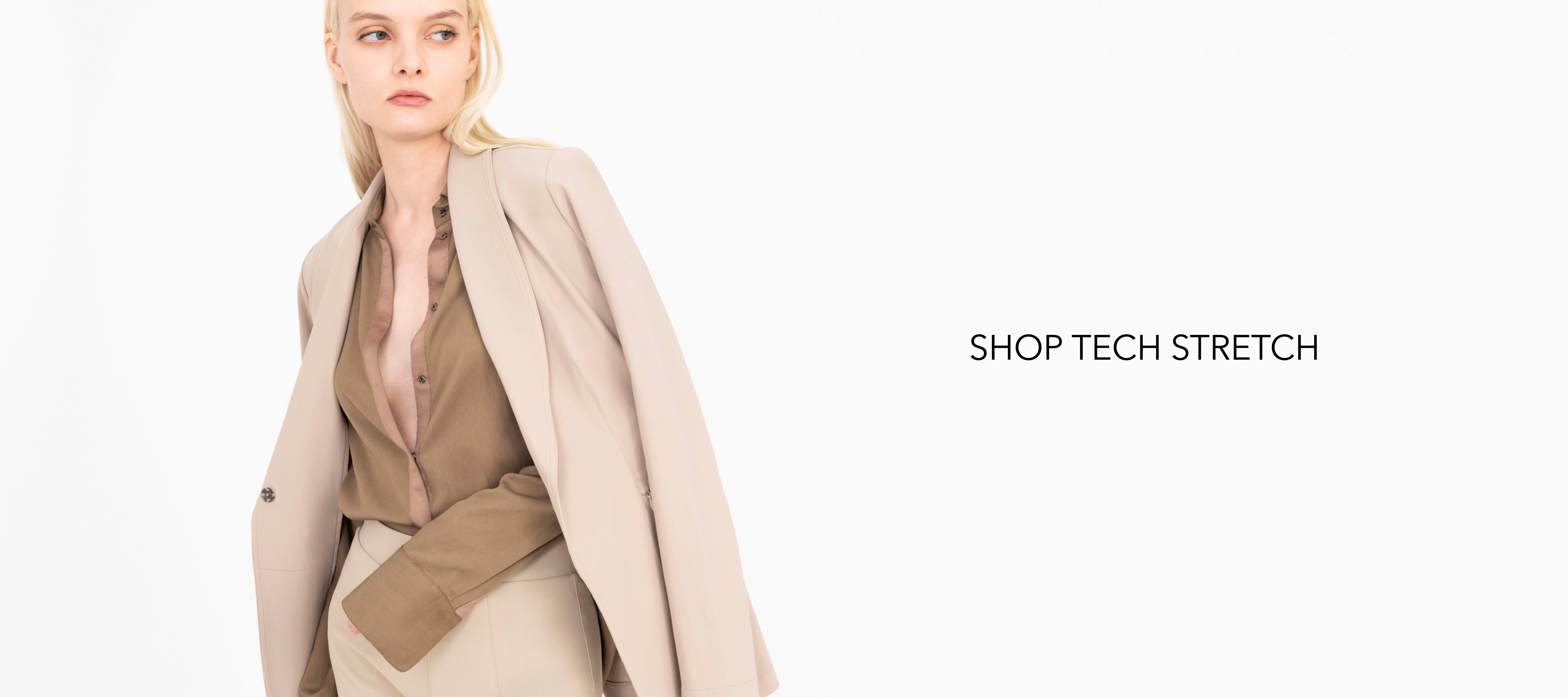 Woman wearing a beige coat over a brown shirt and beige pants on a white background with 'Shop Tech Stretch' text.