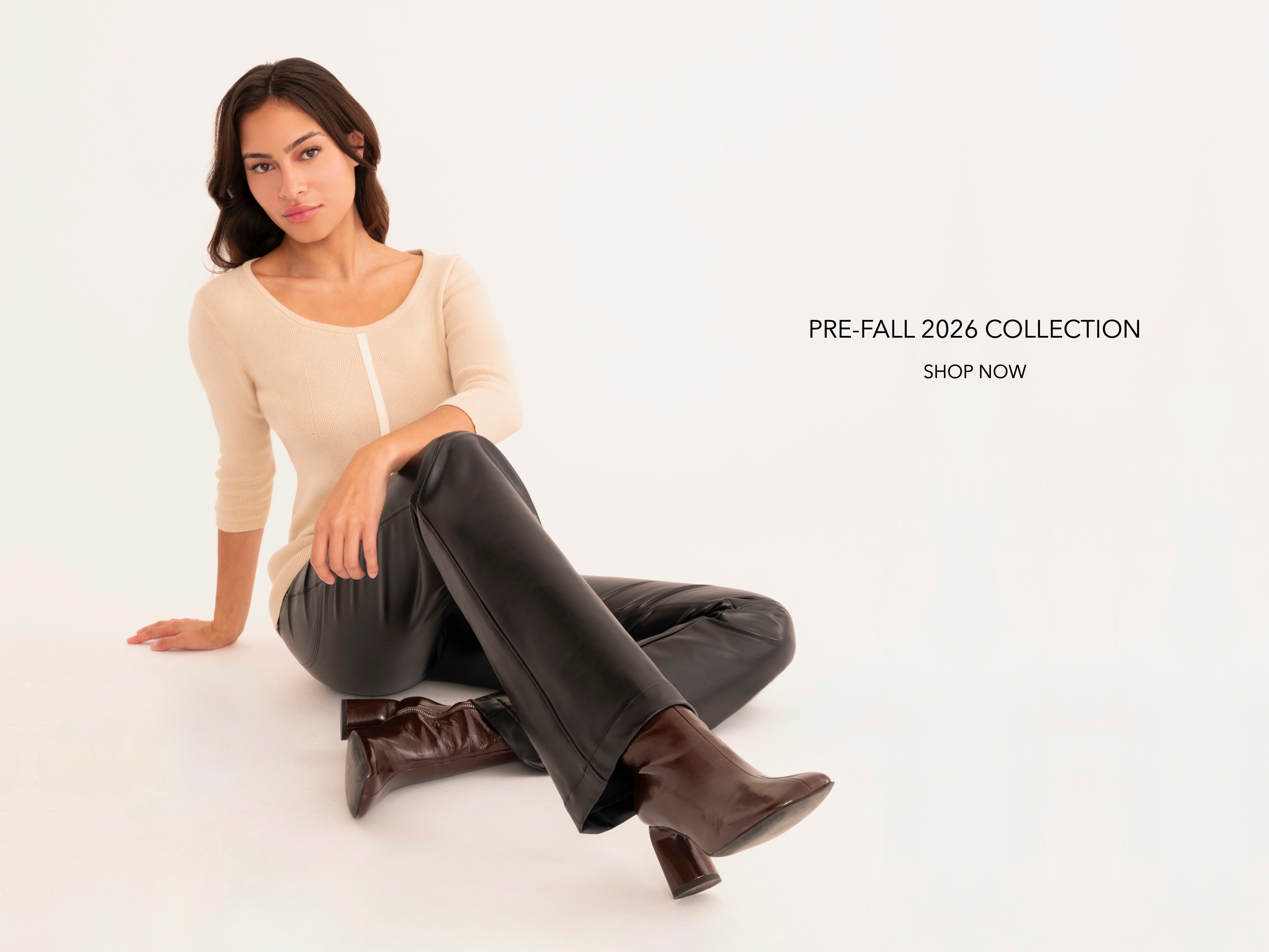 A woman sits on a white studio floor wearing a beige three-quarter-sleeve knit top, black pants, and brown ankle boots. Text on the right reads 'PRE-FALL 2026 COLLECTION SHOP NOW'.