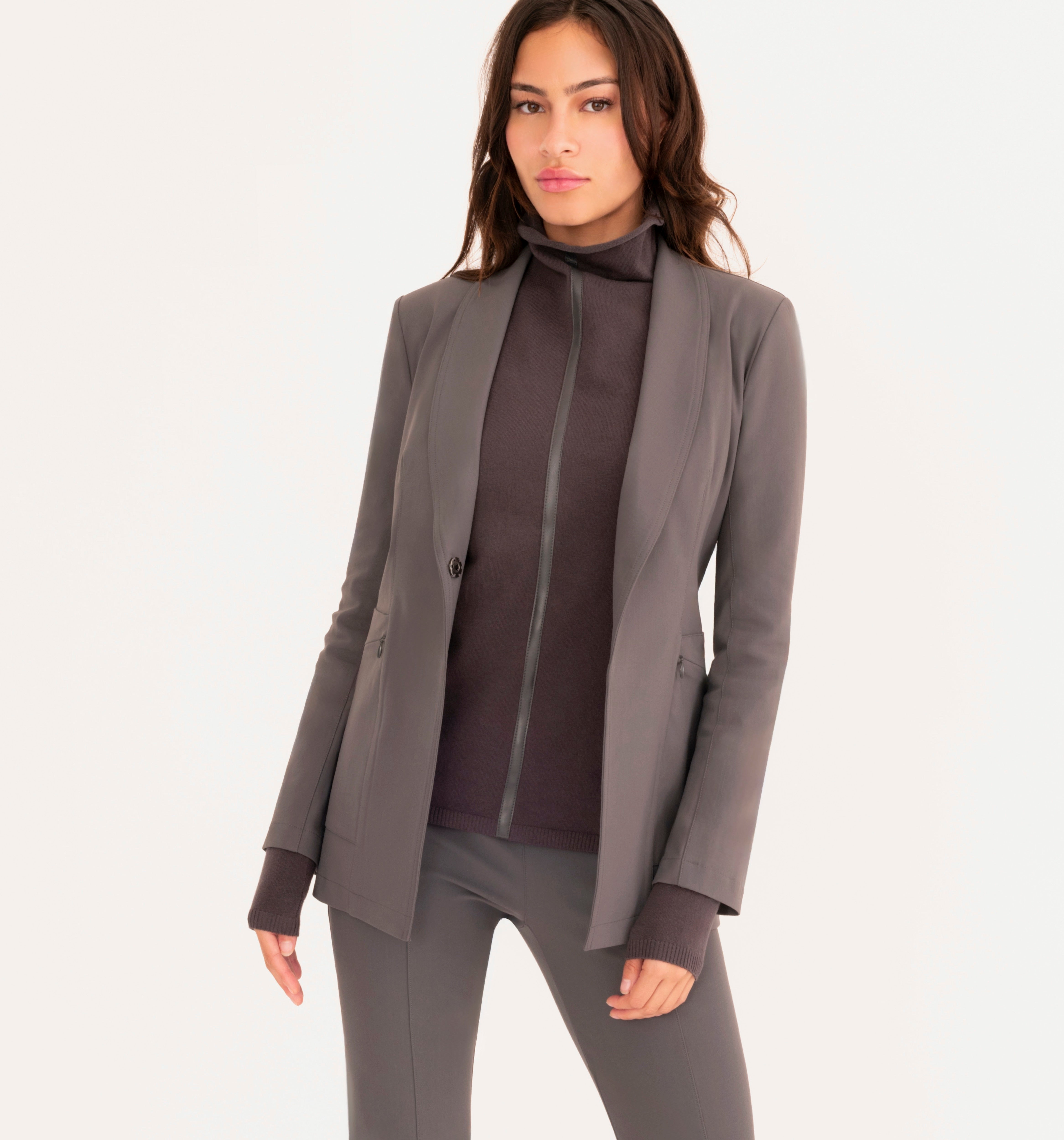 A woman modeling a tailored gray blazer over a gray mock-neck top and matching gray pants, photographed against a clean white studio background.