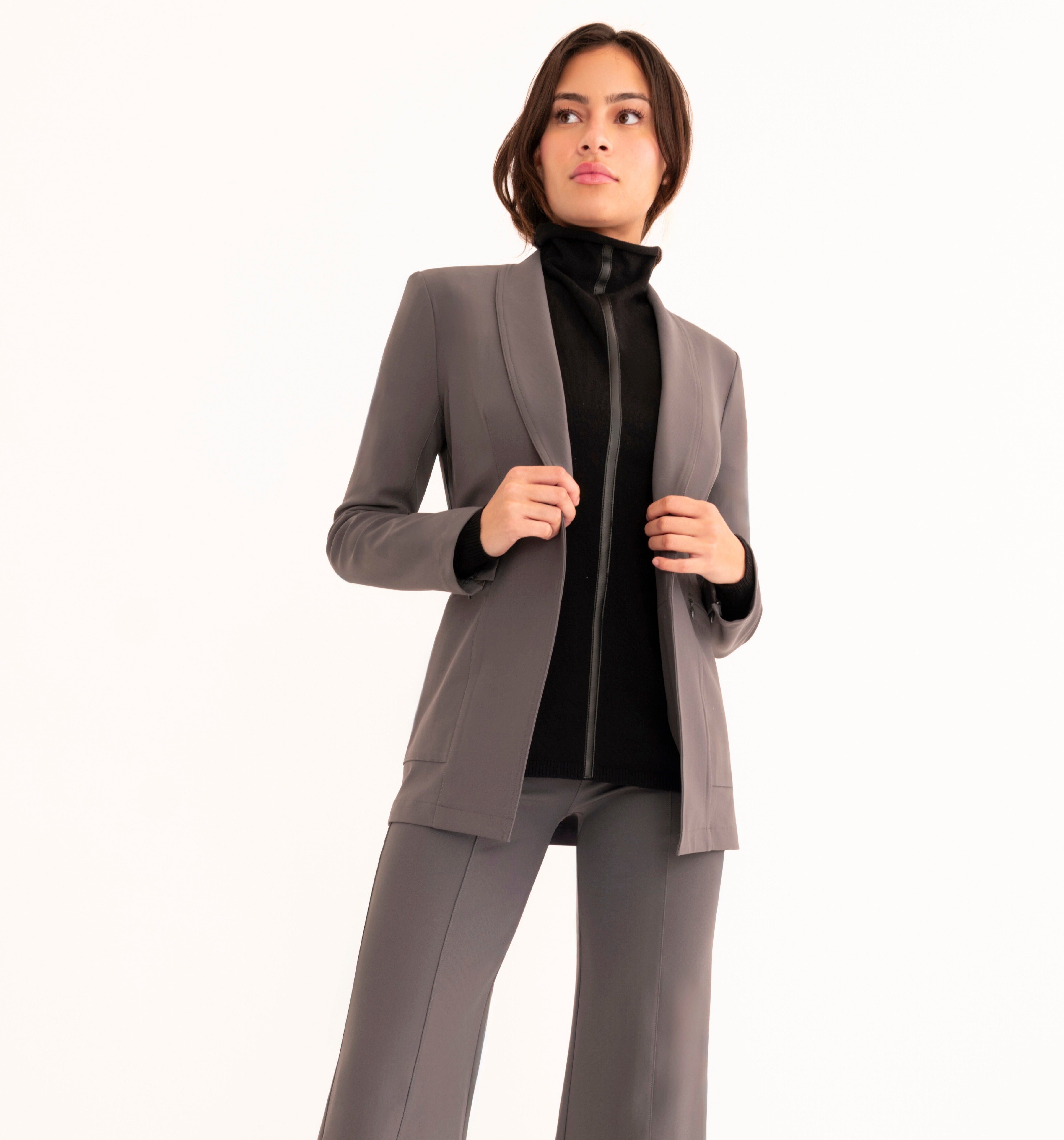 A woman modeling a tailored gray suit with a blazer and matching trousers, worn over a black mock-neck top, against a plain white studio background.