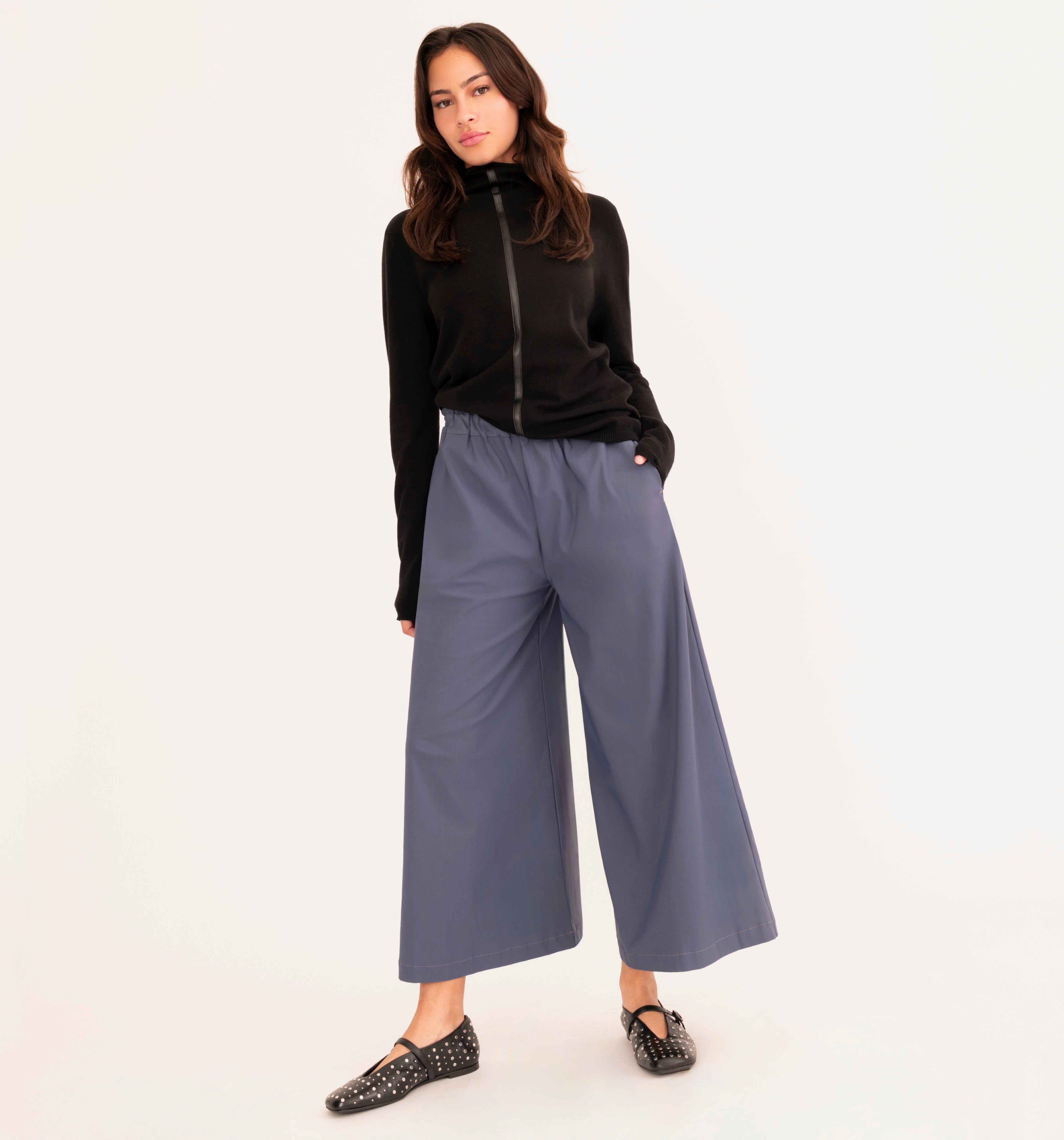 A woman modeling a black mock-neck zip-up top and slate blue wide-leg cropped pants with black studded flats, set against a plain white studio background.