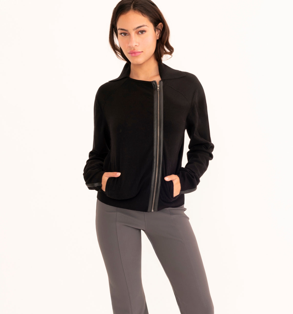 A woman modeling a black zip-up jacket with a stand collar and gray fitted pants, photographed against a clean white studio background.