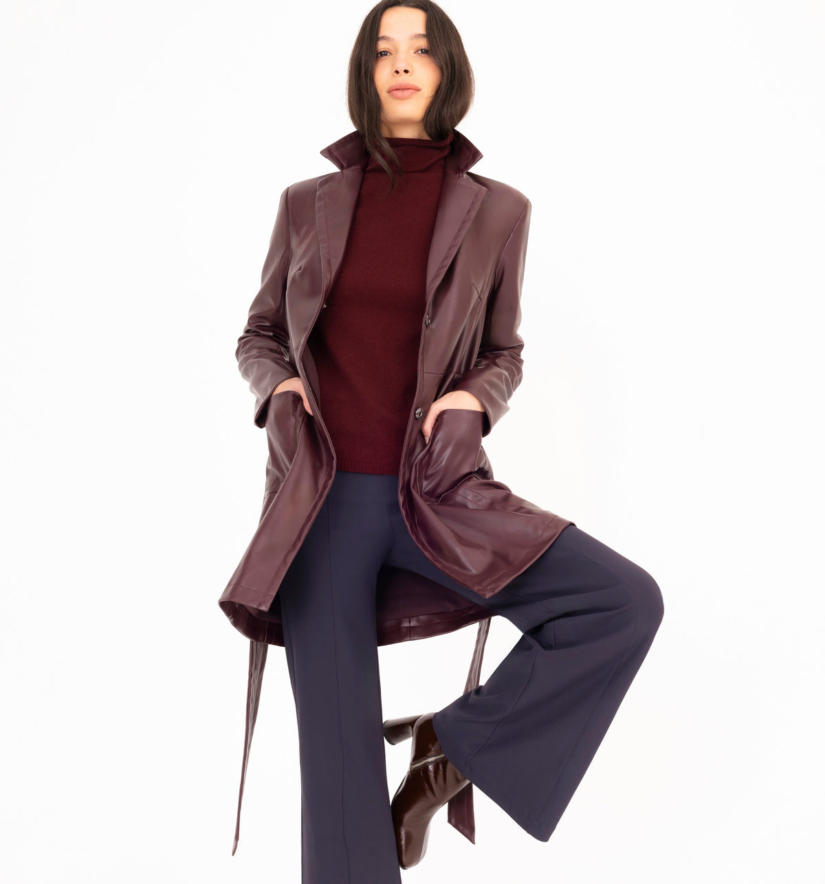 A woman models a long burgundy vegan leather coat over a burgundy turtleneck cashmere sweater and navy wide-leg trousers, posed against a solid white background.