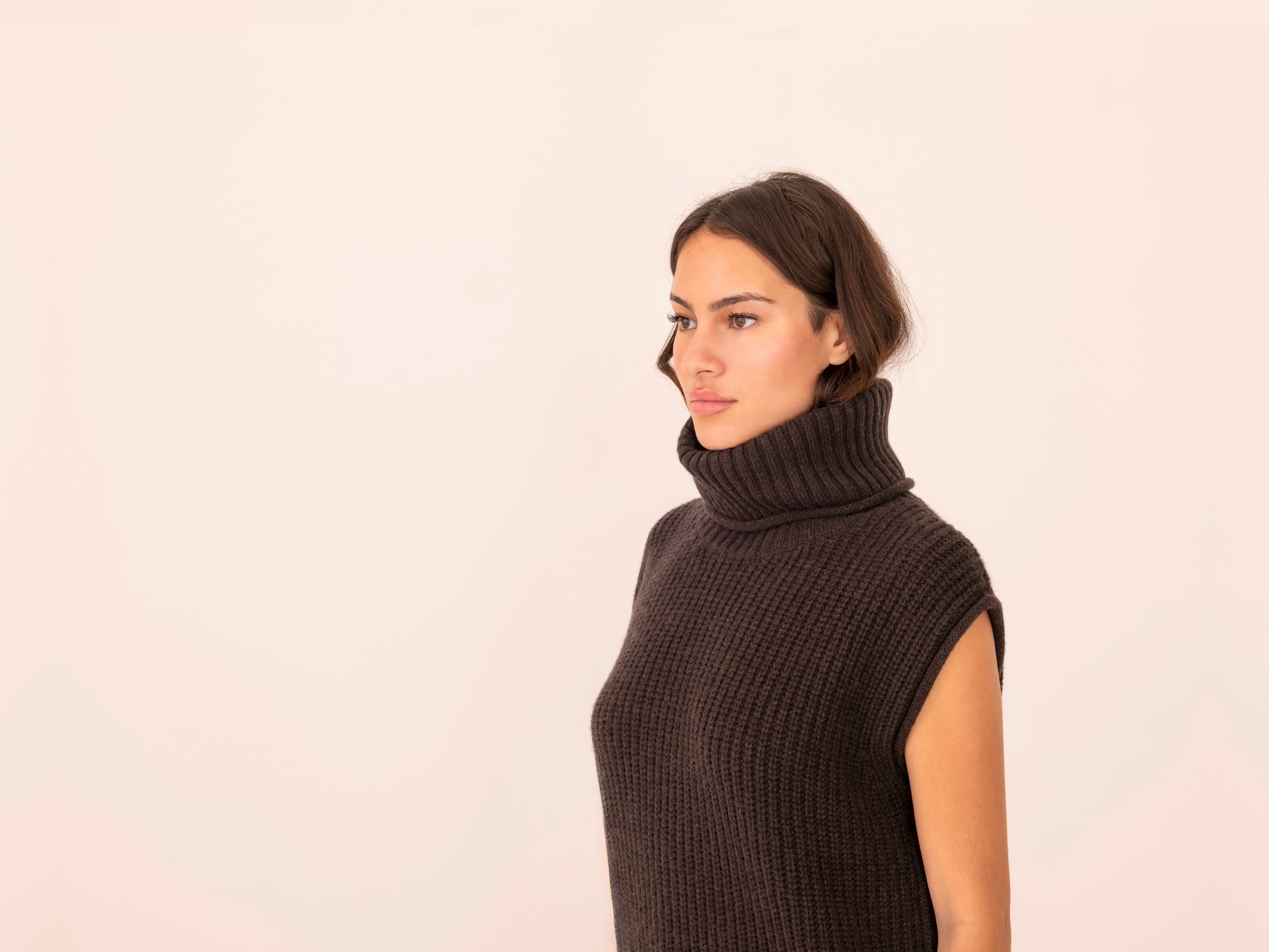 Woman wearing a dark brown sleeveless ribbed knit sweater against a light pink background