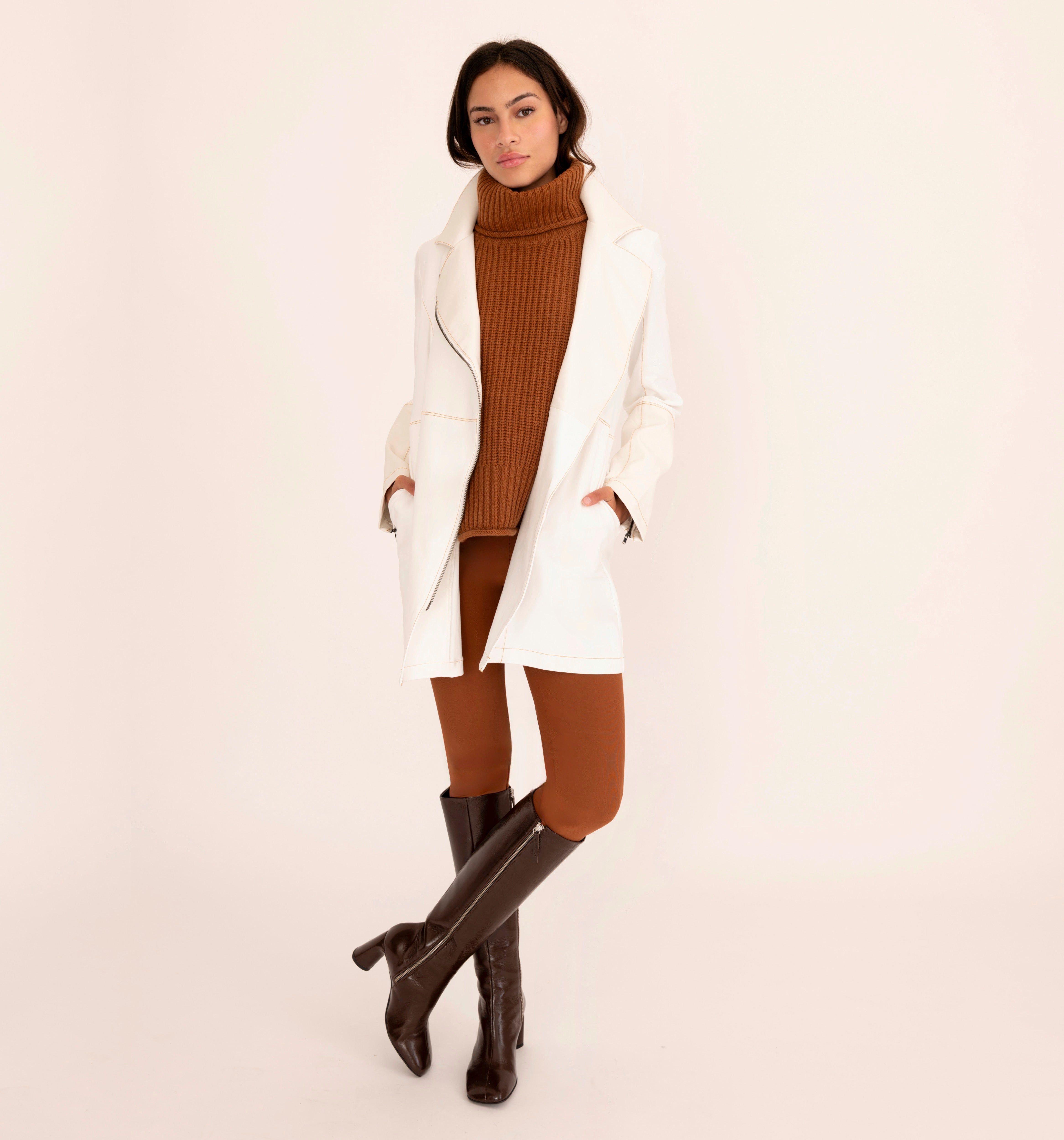 Woman wearing a white coat, brown turtleneck sweater, and knee-high brown boots on a beige background