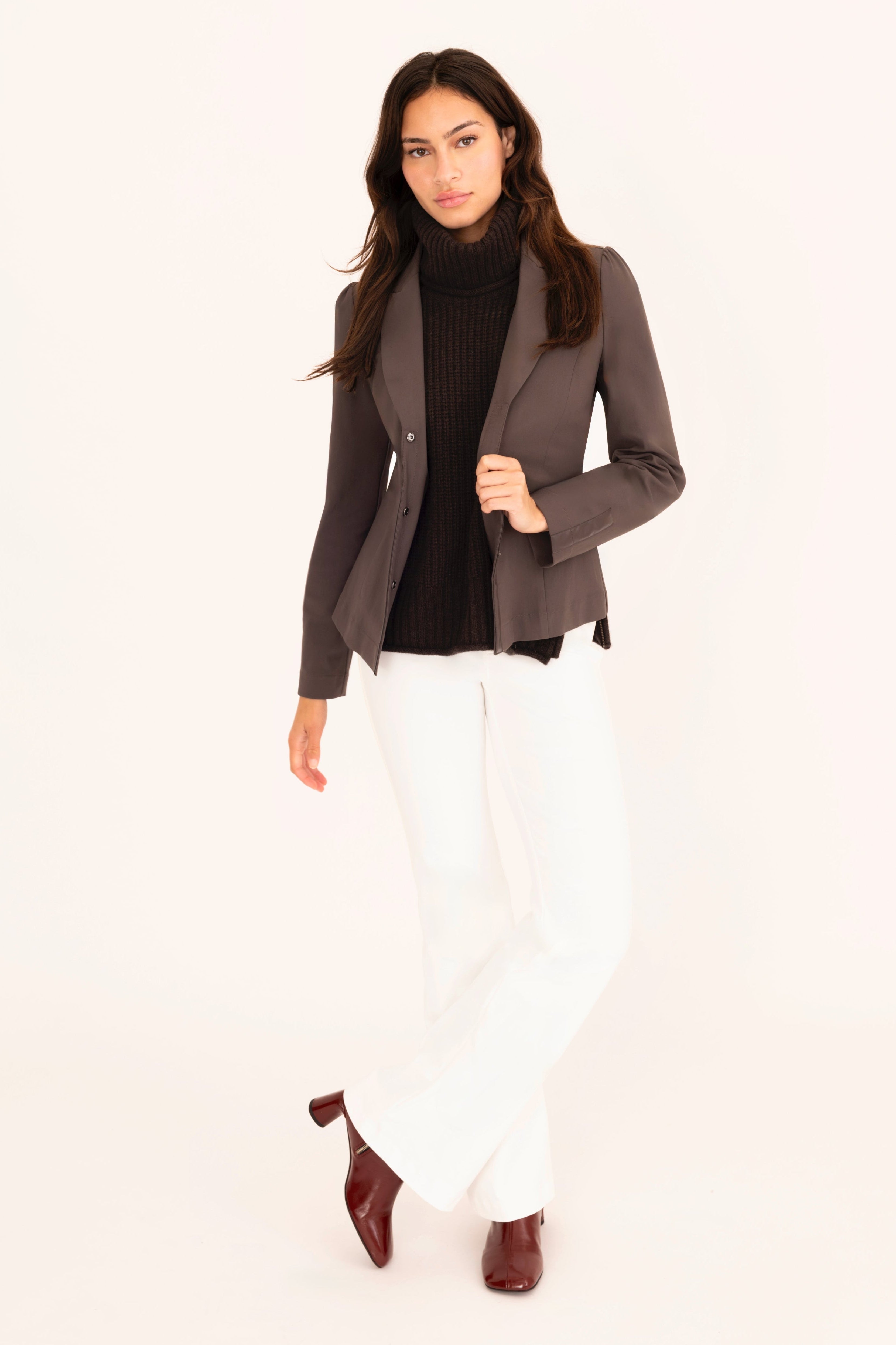 Woman wearing a brown blazer, black turtleneck, white pants, and brown shoes on a white background